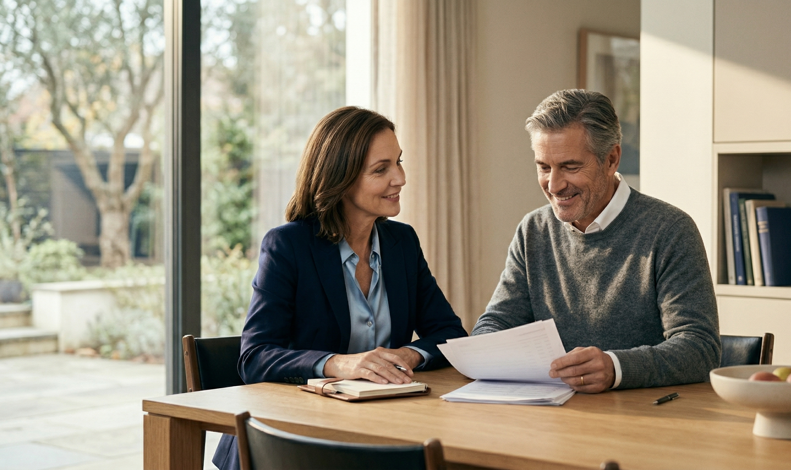 A confident, well-dressed couple in their late 50s sits at a clean, modern dining table reviewing financial documents together.